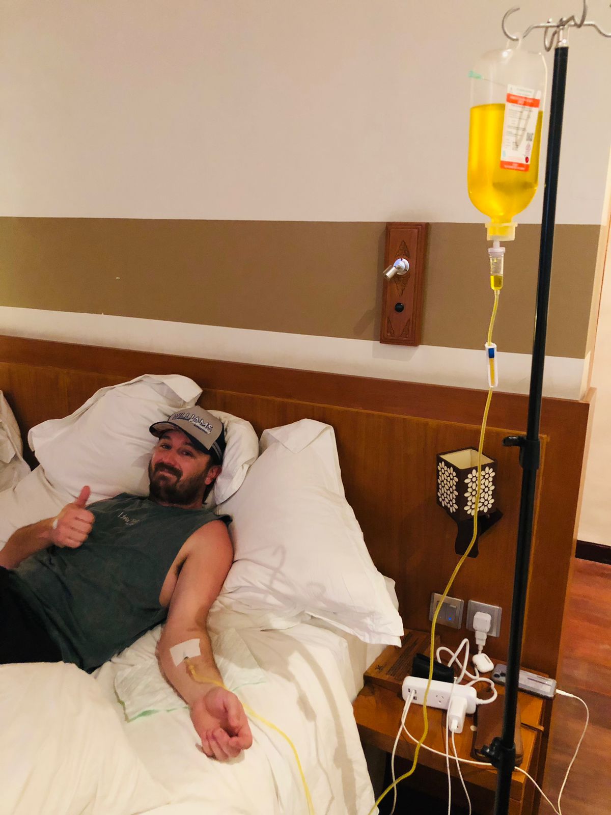 Man smiling and giving a thumbs up while receiving a mobile IV drip treatment in bed.